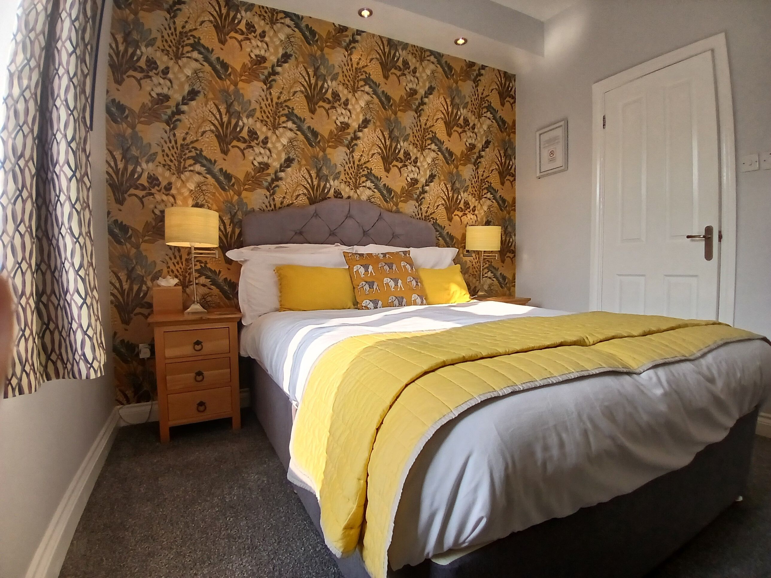 A bedroom with a bed, two bedside tables, a nightstand, a patterned wallpaper, a door, and a wall-mounted light fixture.