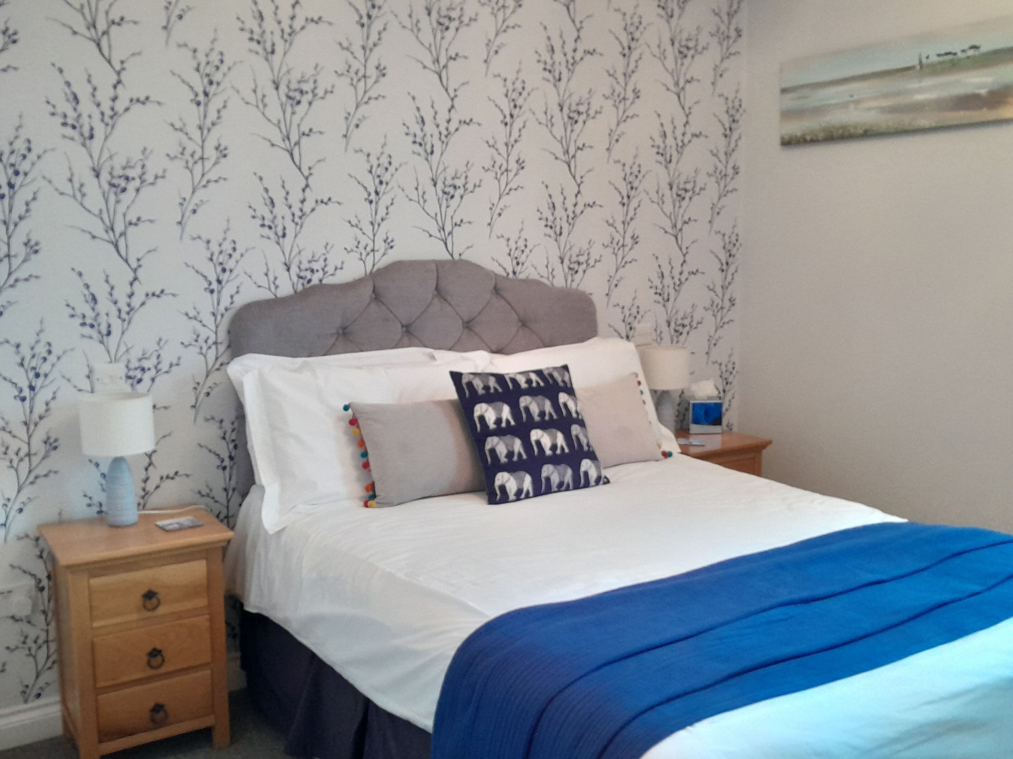 A neatly made bed with a blue blanket, a tufted headboard, and patterned pillows. There are two bedside tables with lamps and a small drawer. The wallpaper has a floral pattern.