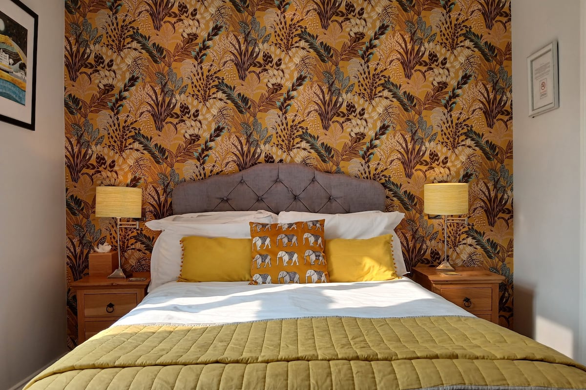 Una cama bien tendida con un colcha amarilla, dos mesitas de noche con lámparas y un papel tapiz floral.