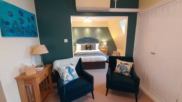 A bedroom with a bed, two armchairs, a nightstand, a lamp, and a painting on the wall.