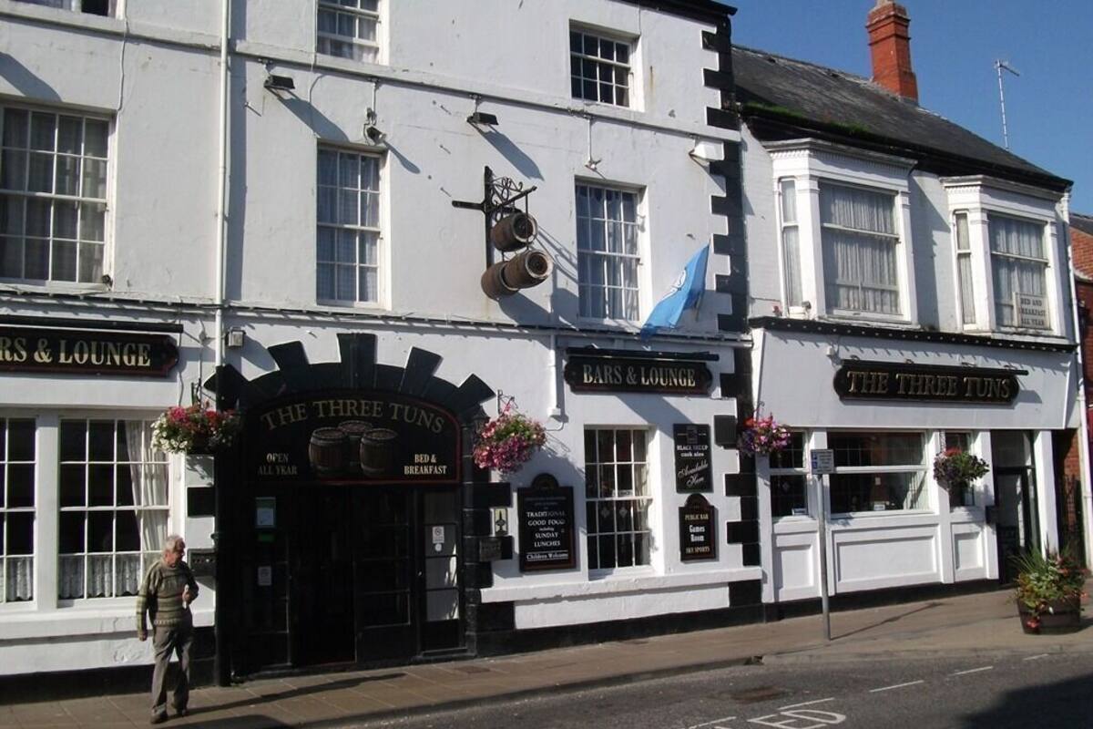 A traditional pub with a sign reading 'Bars & Lounge' and 'The Three Tuns,' featuring a classic white facade with multiple windows and flower arrangements.
