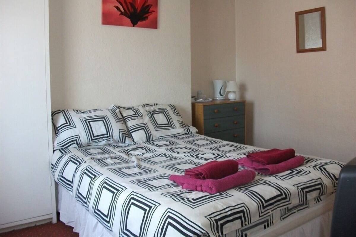 A single bed with a geometric patterned bedspread, a red flower painting on the wall, a small wooden cabinet, and a mirror.