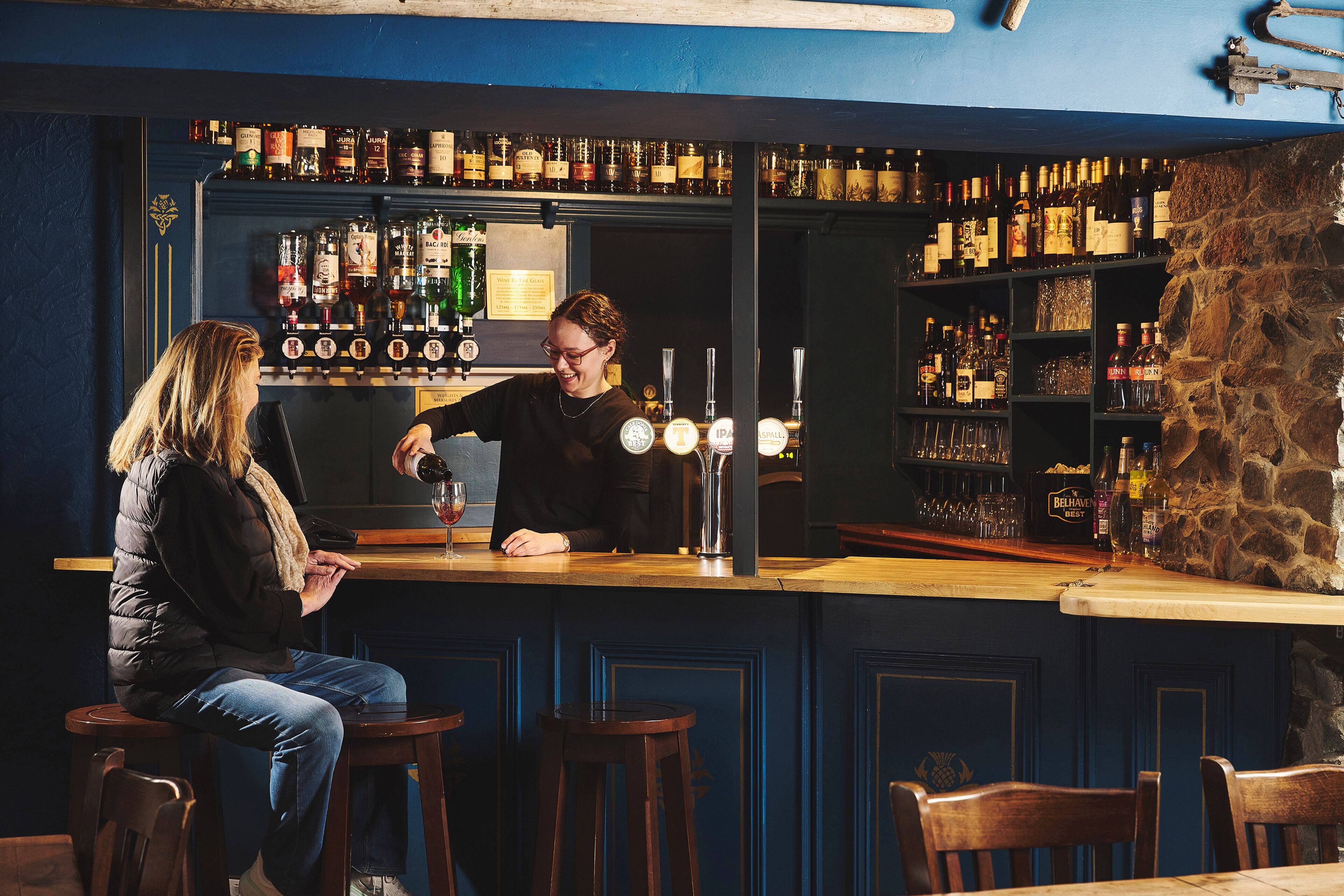 A bar area with a bartender serving a customer. There are shelves with various bottles and glasses behind the bar.