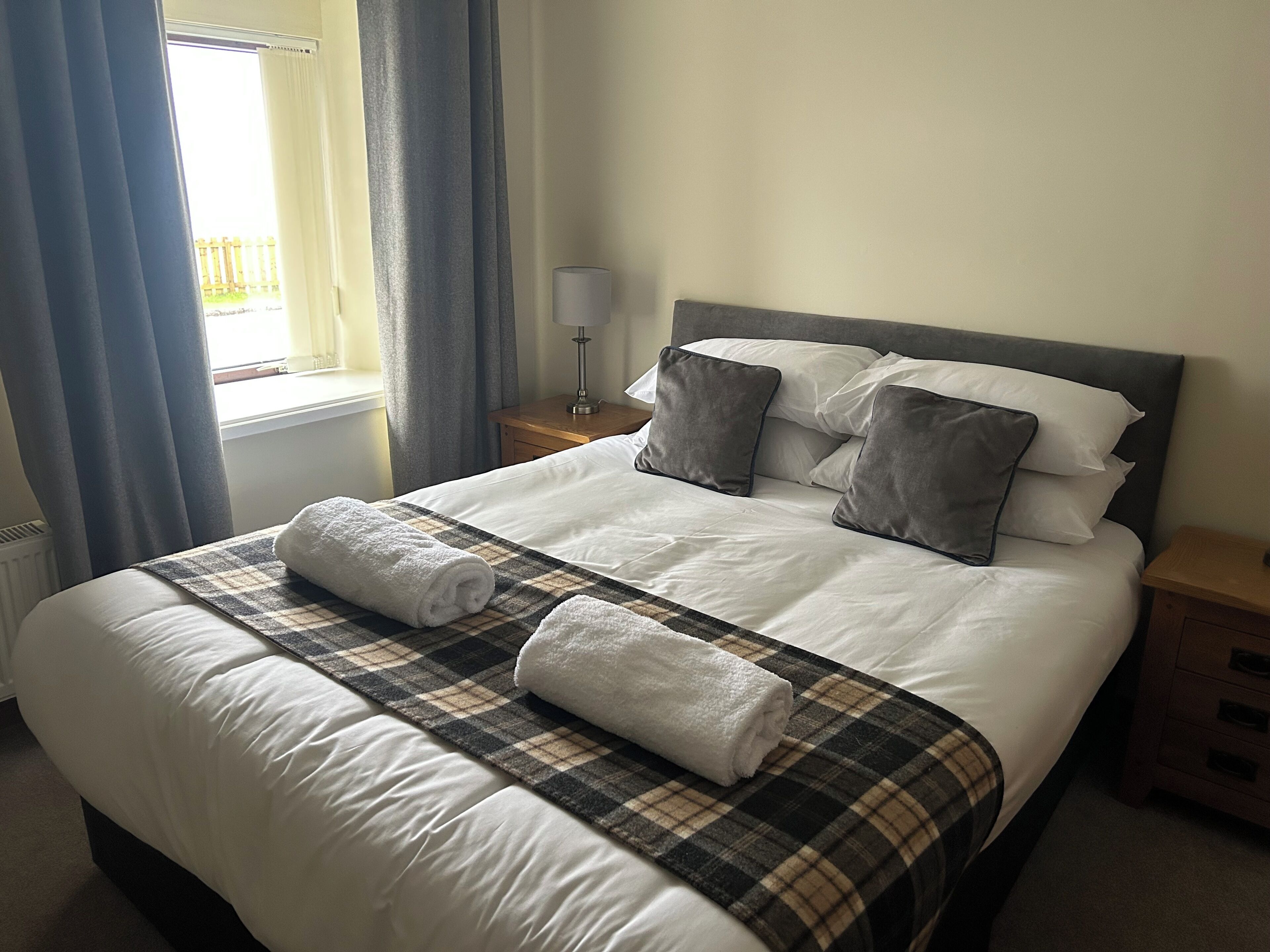 A neatly made bed with a plaid bedspread and two rolled towels, a bedside table with a lamp, and a window with curtains.