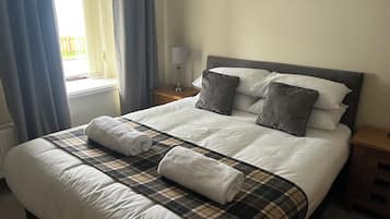 A neatly made bed with a plaid bedspread and two rolled towels, a bedside table with a lamp, and a window with curtains.