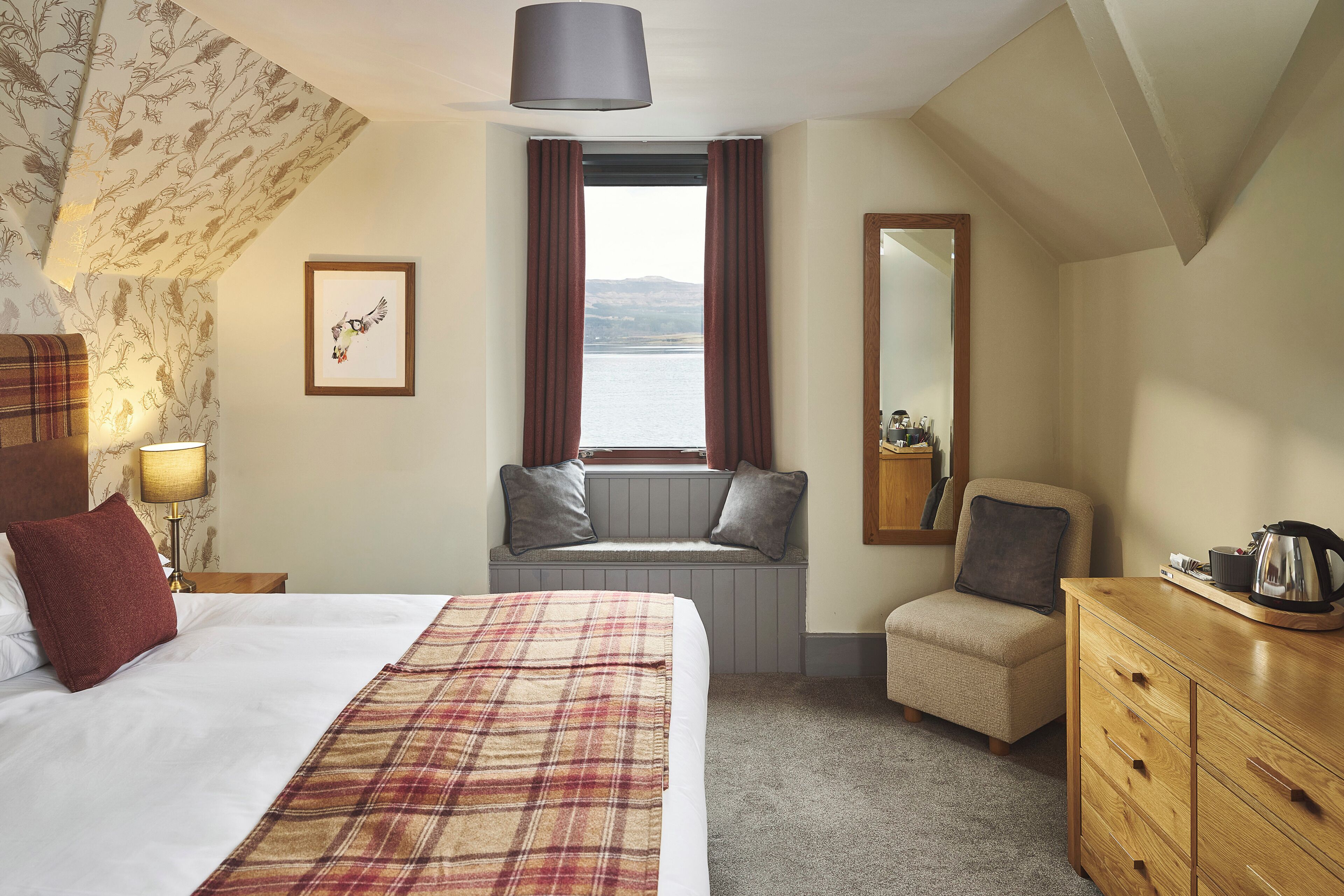 Habitación de hotel con cama, ventana con cortinas, zona de estar con vistas, cómoda y lámpara.