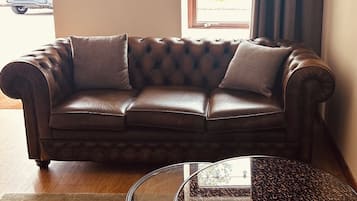 Een bruine leren bank met een glazen salontafel voor een raam met gordijnen.