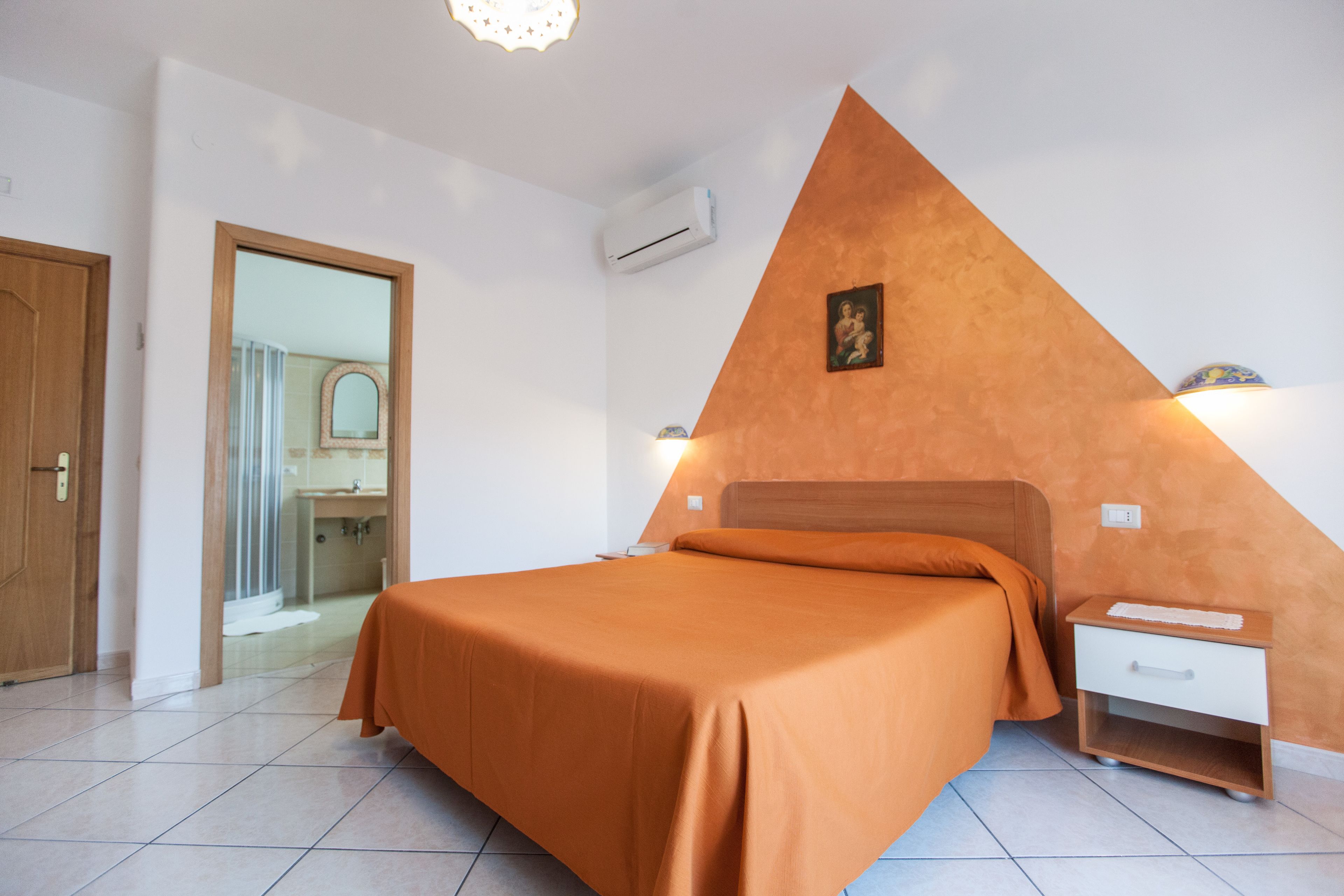 A bedroom with a bed, a nightstand, a wall-mounted air conditioner, and a bathroom visible through an open door.