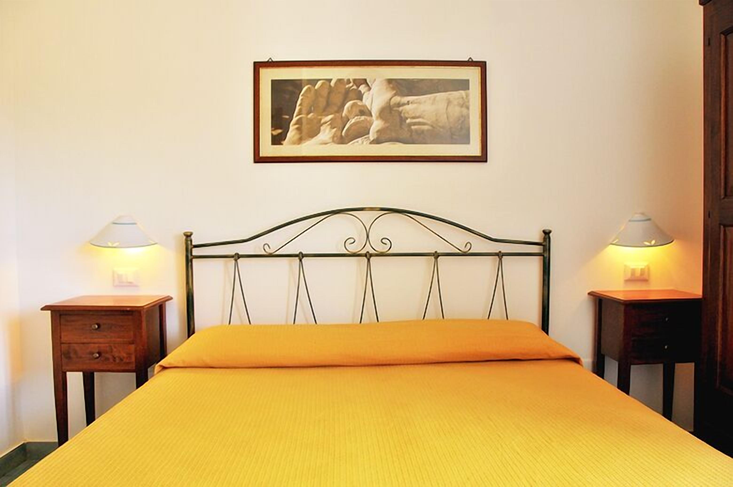 Uma cama com cabeceira de metal, duas mesas de cabeceira com abajures, um quadro na parede e uma parede lisa.
