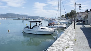 Un port de plaisance où des bateaux sont amarrés le long d’une promenade pavée, avec des bâtiments et un ciel dégagé en arrière-plan.