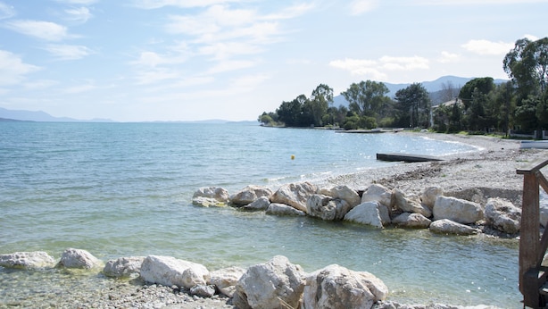 Un rivage rocheux bordé d’une eau claire, une jetée en bois et, au loin, une colline boisée.