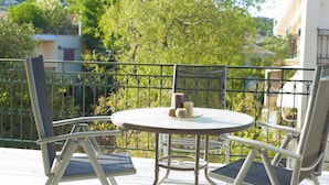 Un balcon avec une table ronde et deux chaises, une balustrade et une vue sur les arbres et les bâtiments en arrière-plan.