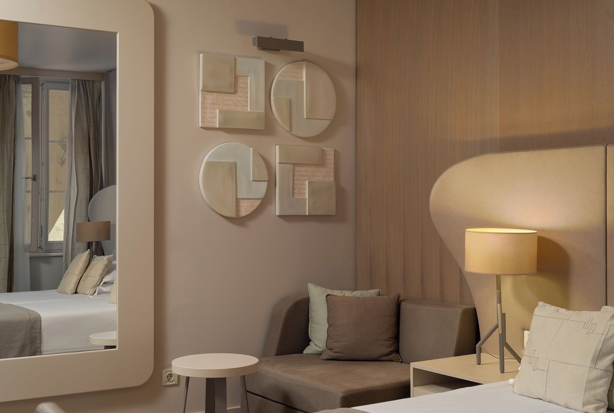 A modern hotel room with a bed, a sofa, a small table, and a lamp. There are abstract wall art pieces and a mirror.
