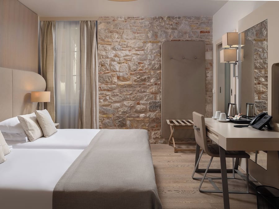 Quarto de hotel com uma cama grande, uma escrivaninha, uma cadeira, chuveiro e parede de pedra.
