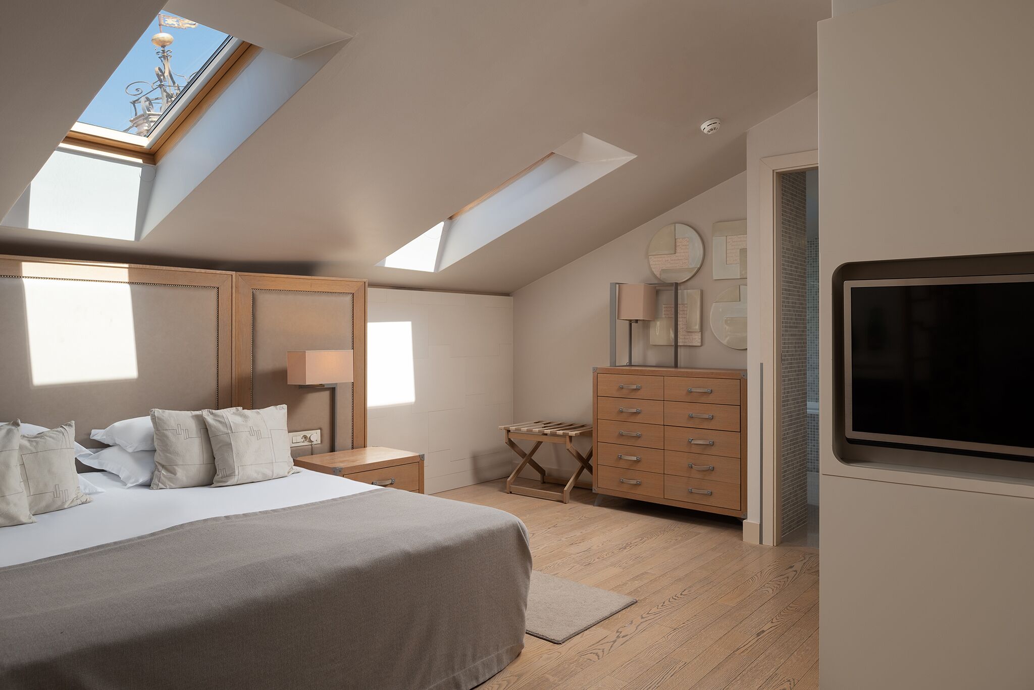 Un dormitorio con tragaluces, una cama con almohadas, un aparador, un televisor y una lámpara.