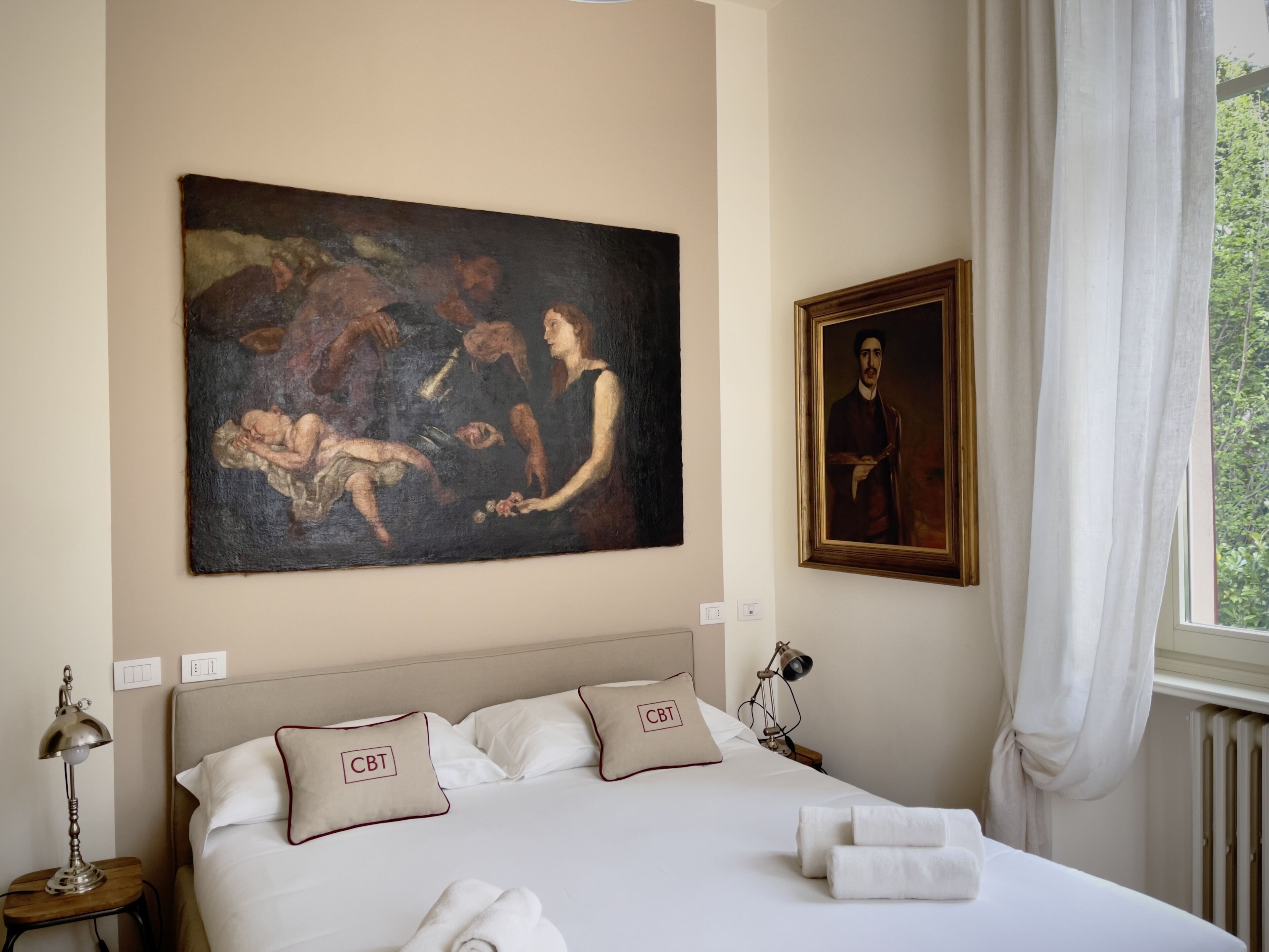Una camera d'albergo con un quadro di grandi dimensioni appeso al muro, un letto rifatto con lenzuola bianche e due cuscini con la scritta "CBT", una lampada da comodino e una finestra con tende.