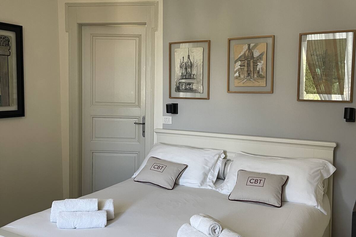 A neatly made bed with white linens and two pillows with 'CBT' embroidered on them, a door to the left, and three framed pictures on the wall.