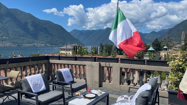 A terrace with outdoor seating, a flag, and a view of mountains and a lake.