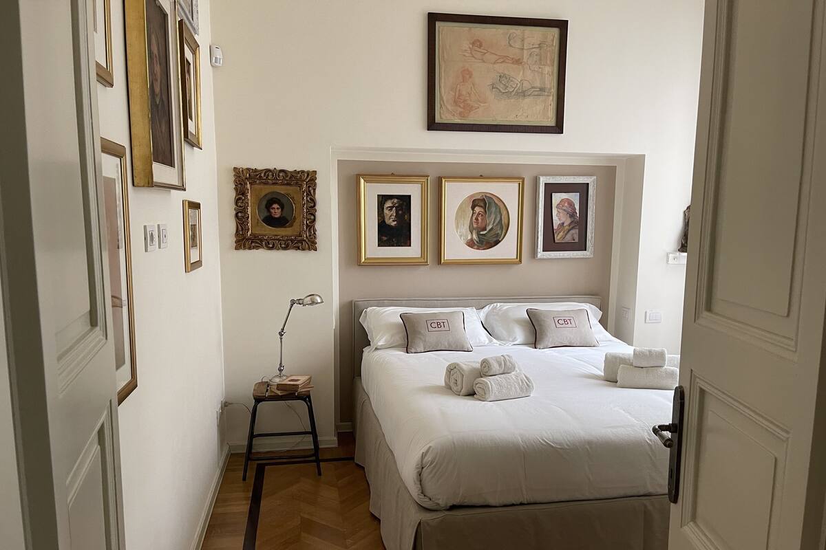 A bedroom with a large bed, a nightstand with a lamp, and several framed pictures on the wall.