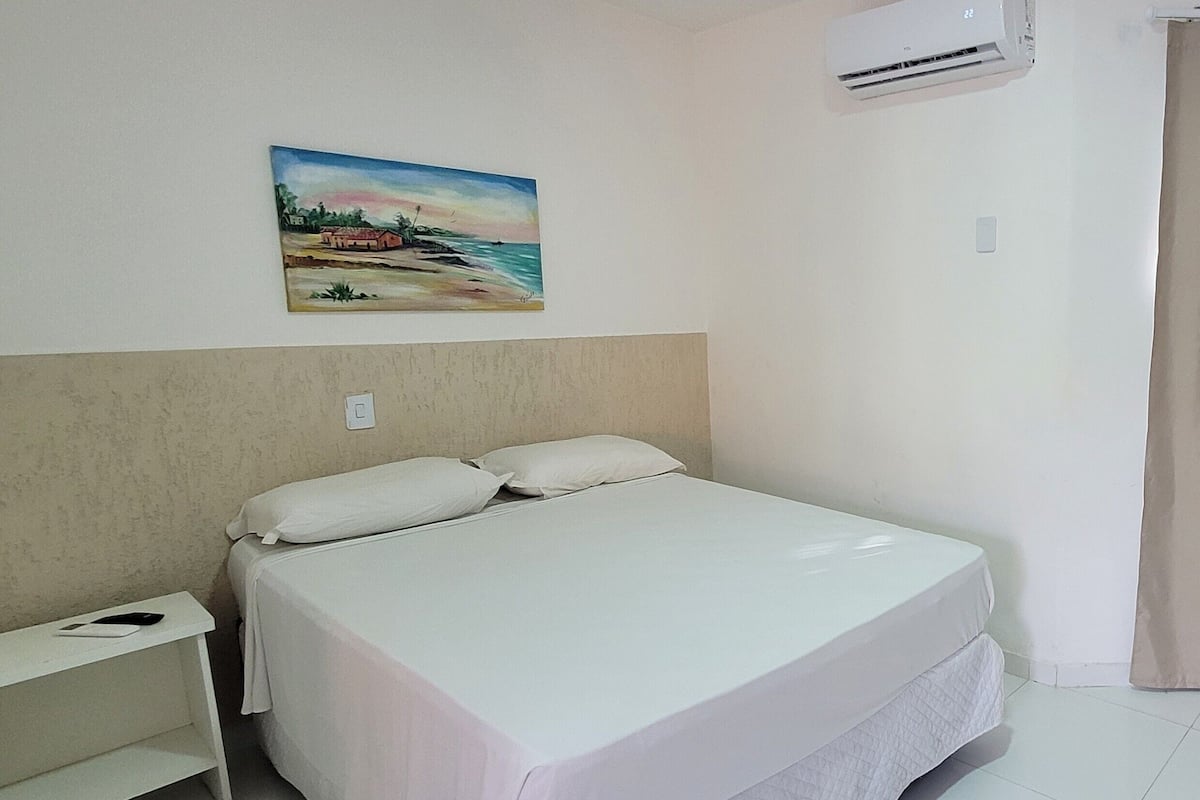 A hotel room with a bed, a painting on the wall, an air conditioner, a curtain, and a mirror.