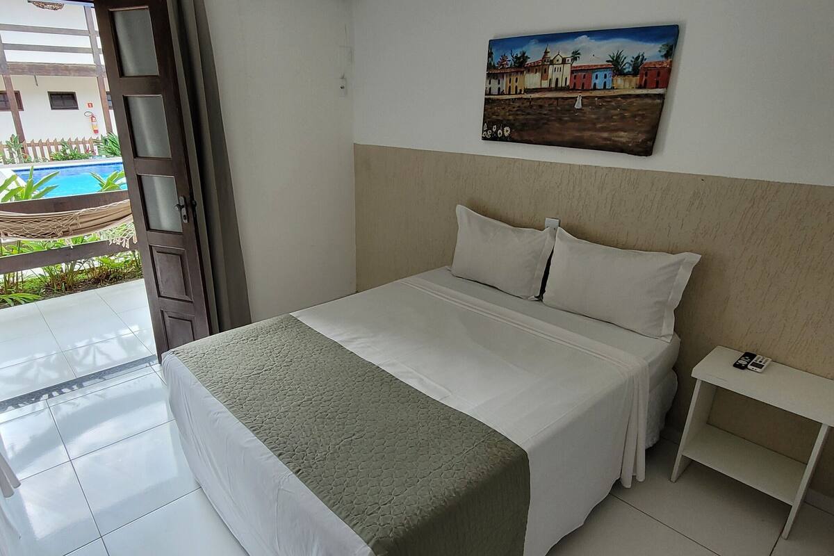 Habitación de hotel con una cama, una mesita de noche con teléfono, un cuadro en la pared y vistas a una piscina exterior.