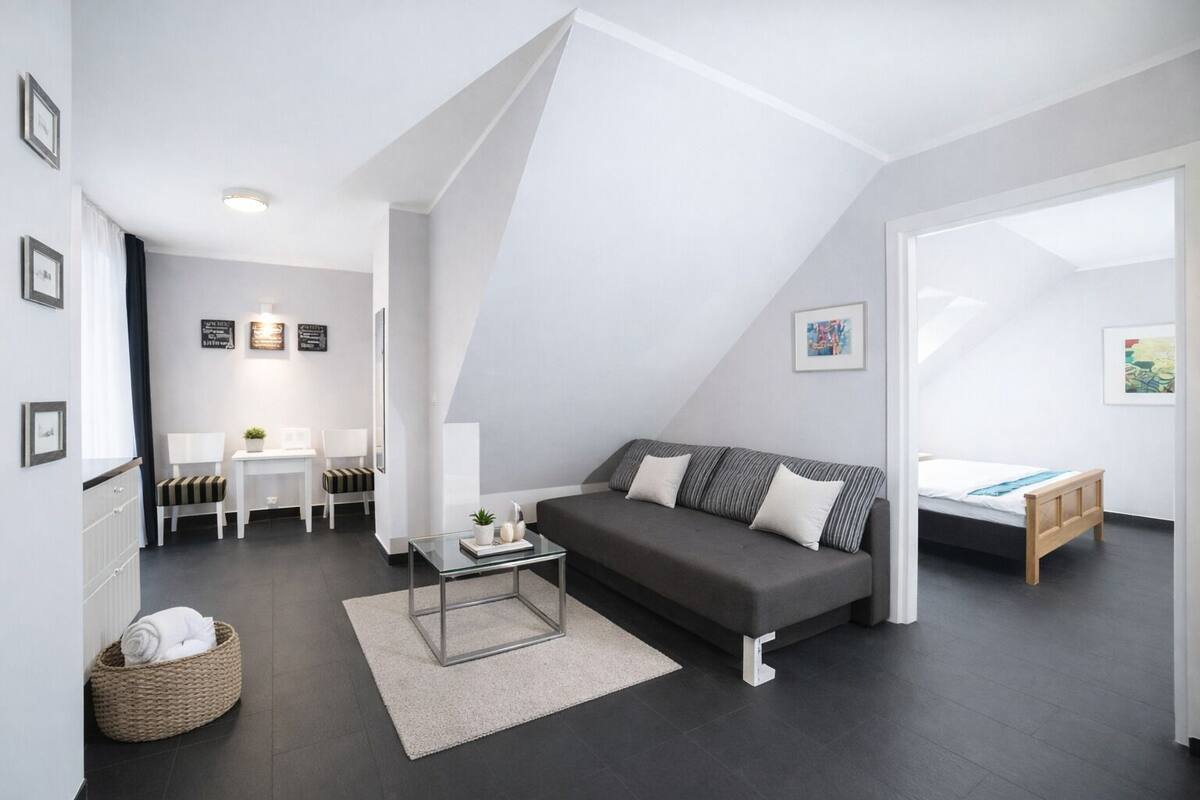 A modern, well-lit loft bedroom with a dark gray sofa, a small glass coffee table, and a wooden bed.