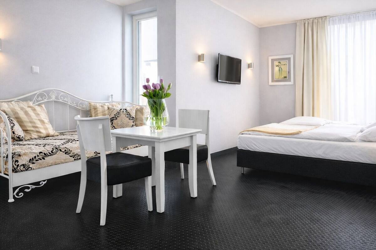 A modern hotel room with a white bed, a white sofa, a small table, and a TV mounted on the wall.