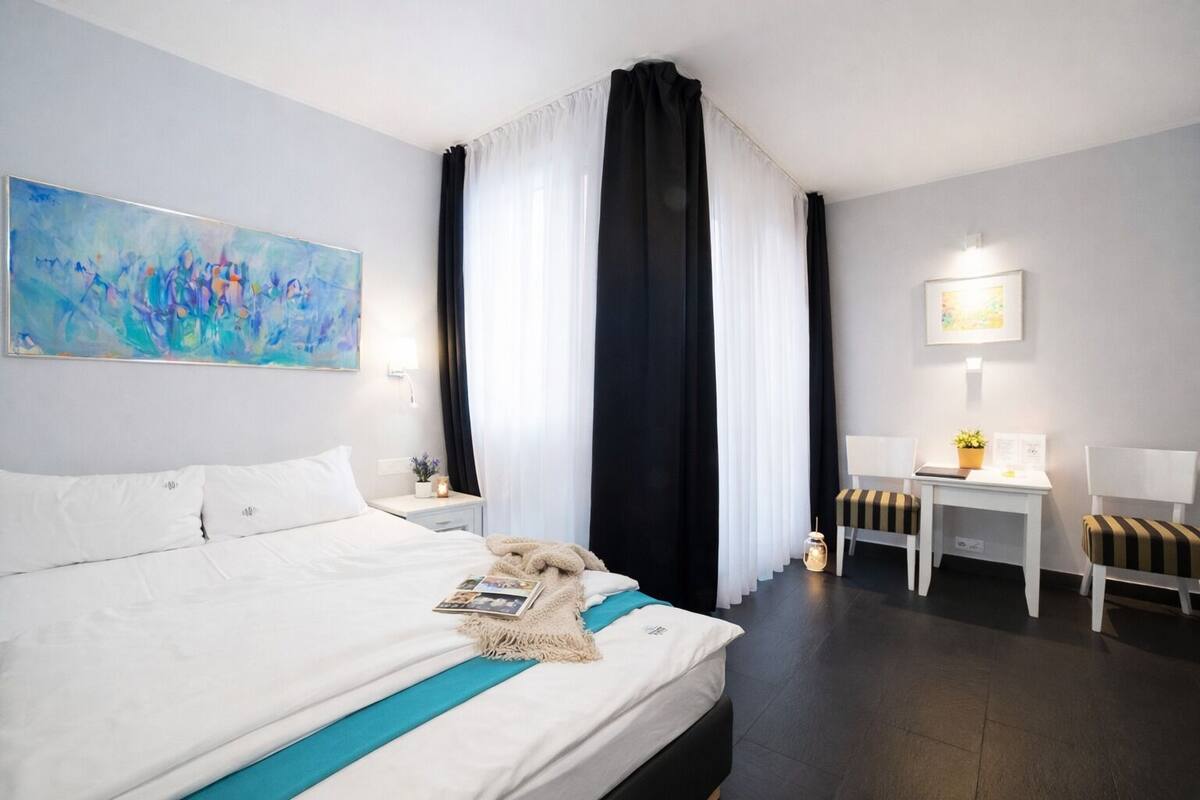 A hotel room with a large bed, a desk with two chairs, a small table with a plant, and a painting on the wall.