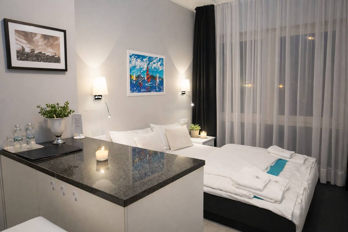 A modern hotel room with a bed, a desk, and a nightstand. There are two wall-mounted lamps, a framed picture, and a potted plant on the desk.