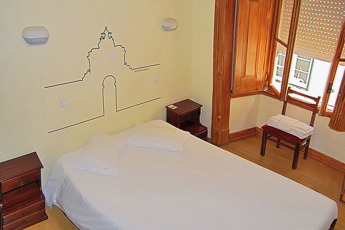 Un dormitorio con cama, mesita de noche, una silla de madera, ventana con persianas y un mapa mural.