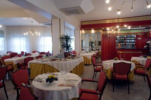Un espace repas avec des tables rondes dressées pour le service, des chaises à l’assise rembourrée de couleur rouge et un bar avec des étagères remplies de bouteilles.