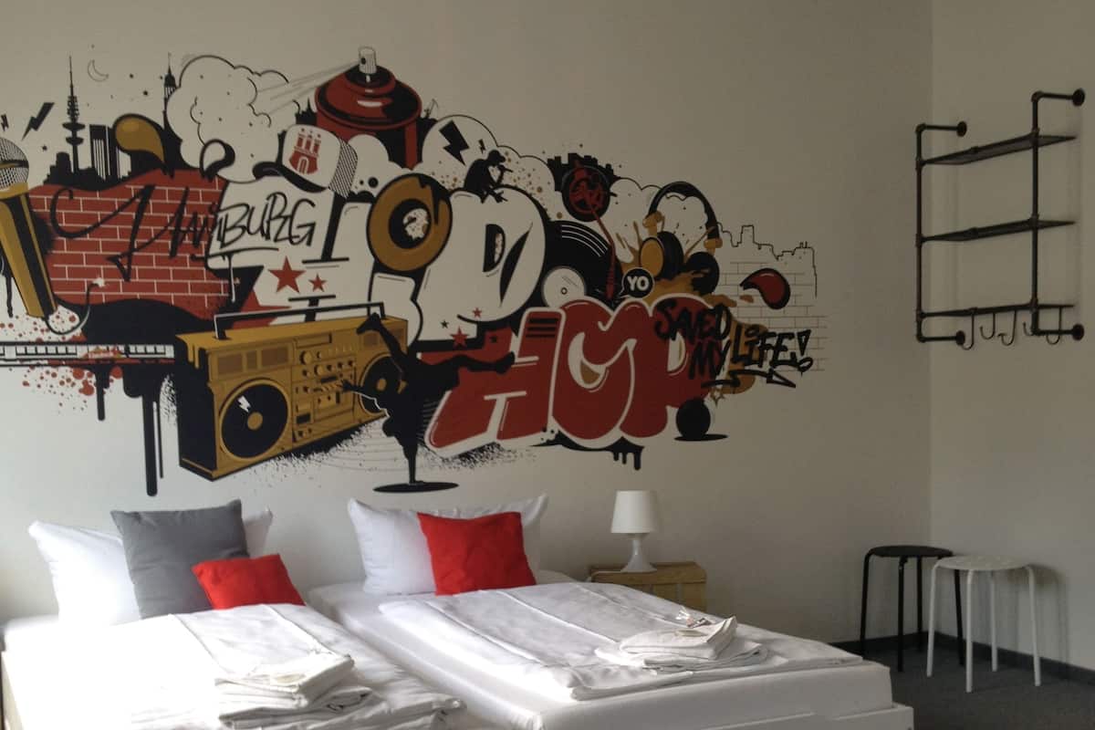 A modern bedroom with a large bed, white bedding, red and gray pillows, a wall mural, and a small table with a lamp.