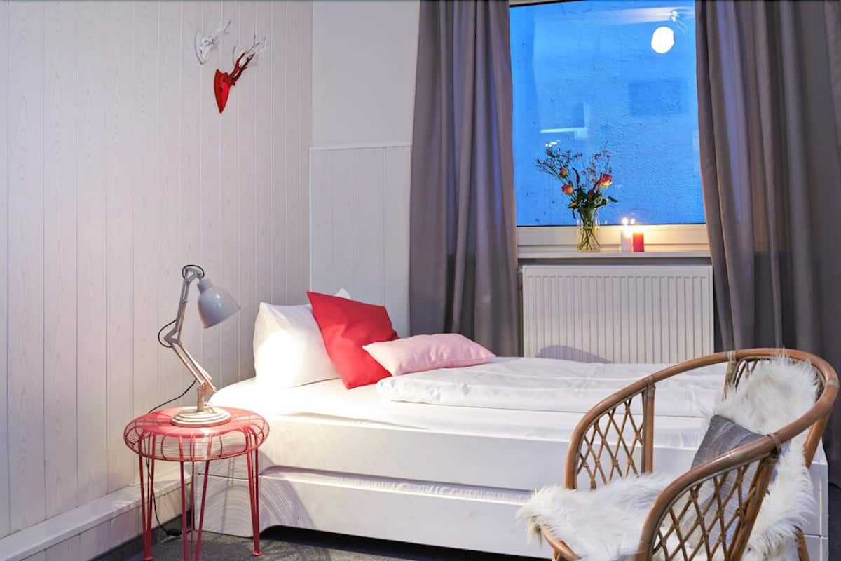 A bedroom with a bed, a chair, a lamp, a window with curtains, and a vase with flowers on the windowsill.