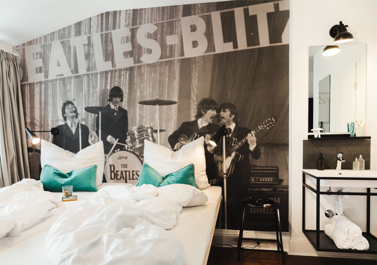 Hotelkamer met een muurschildering met Beatles-thema, een bed met witte beddengoed, een nachtkastje met een lamp en een badkamer met een spiegel en toiletartikelen.