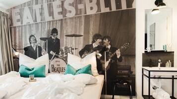 Ein Hotelzimmer mit Wandgemälde zum Thema Beatles, einem Bett mit weißer Bettwäsche, einem Nachttisch mit Lampe und einem Badezimmer mit Spiegel und Pflegeprodukten.