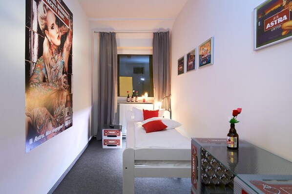 Una habitación de hotel con una cama, una mesita con una botella, un cartel en la pared y una ventana con cortinas.