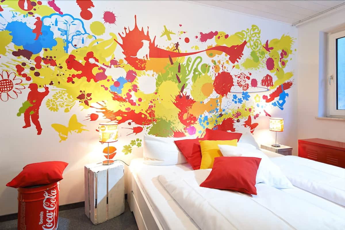 A hotel room with a colorful abstract mural on the wall, a bed with white bedding and red pillows, a nightstand with a lamp, and a Coca-Cola can on the floor.