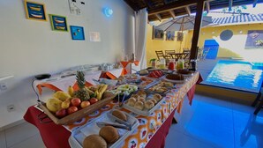 Una mesa de buffet con diversas frutas y pasteles, una zona de piscina visible a través de la ventana, y un techo con vigas a la vista.