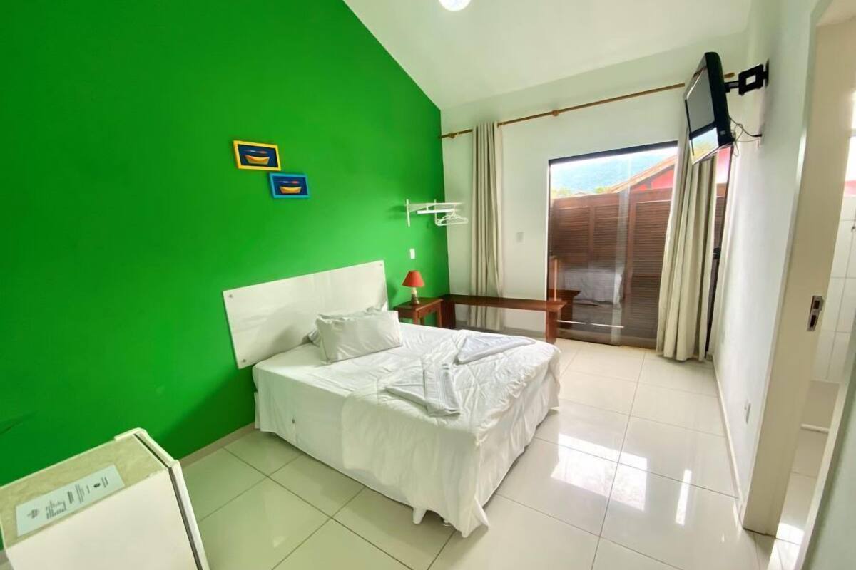 A bedroom with a green wall, a bed with white bedding, a small table with a lamp, and a sliding glass door leading to an outdoor area.
