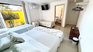 Habitación de hotel con dos camas, televisor, kitchenette pequeña y balcón con paredes amarillas y plantas.