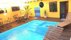 Una piscina con sillones de madera y una mesa, rodeada de paredes amarillas y plantas en macetas.