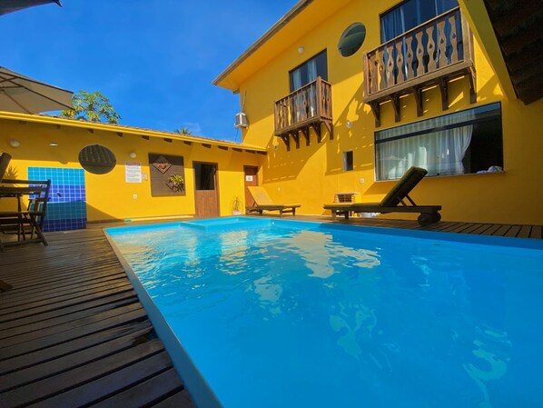 A swimming pool area with a wooden deck, lounge chairs, and a yellow building with balconies.