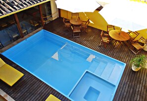 A blue swimming pool with a diving board, surrounded by wooden deck and yellow chairs.