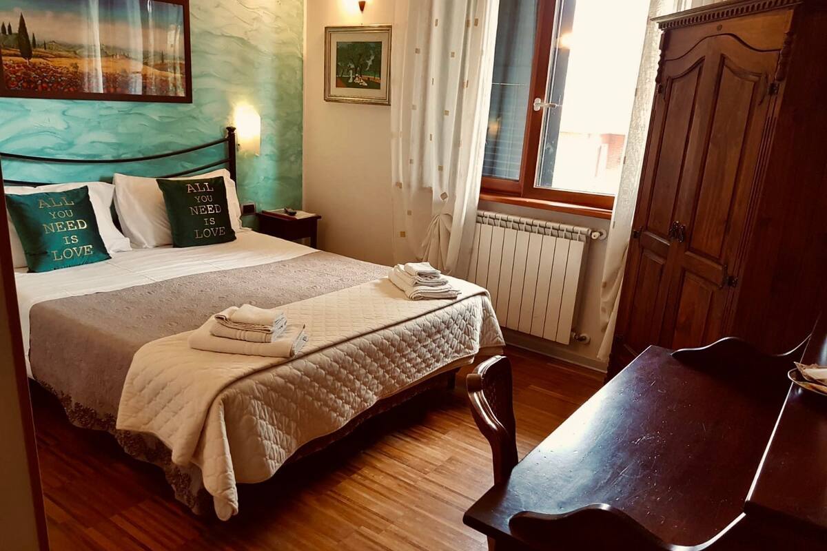 Un lit bien fait, avec des oreillers, une table de chevet avec une lampe, une chaise en bois et une vue sur l’extérieur par une fenêtre.