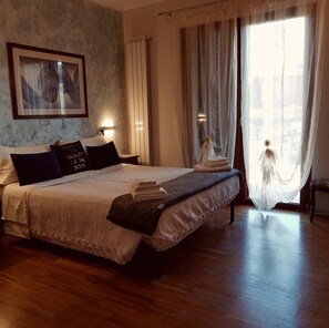 A neatly made bed with a white comforter and dark pillows, a bedside lamp, a framed picture on the wall, and a window with sheer curtains. - Aurelia Garden Gold Bed & Breakfast (Rome)