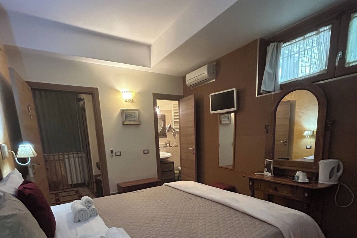 Une chambre d’hôtel avec un grand lit, une table de chevet avec une lampe, un miroir et une fenêtre avec des rideaux.