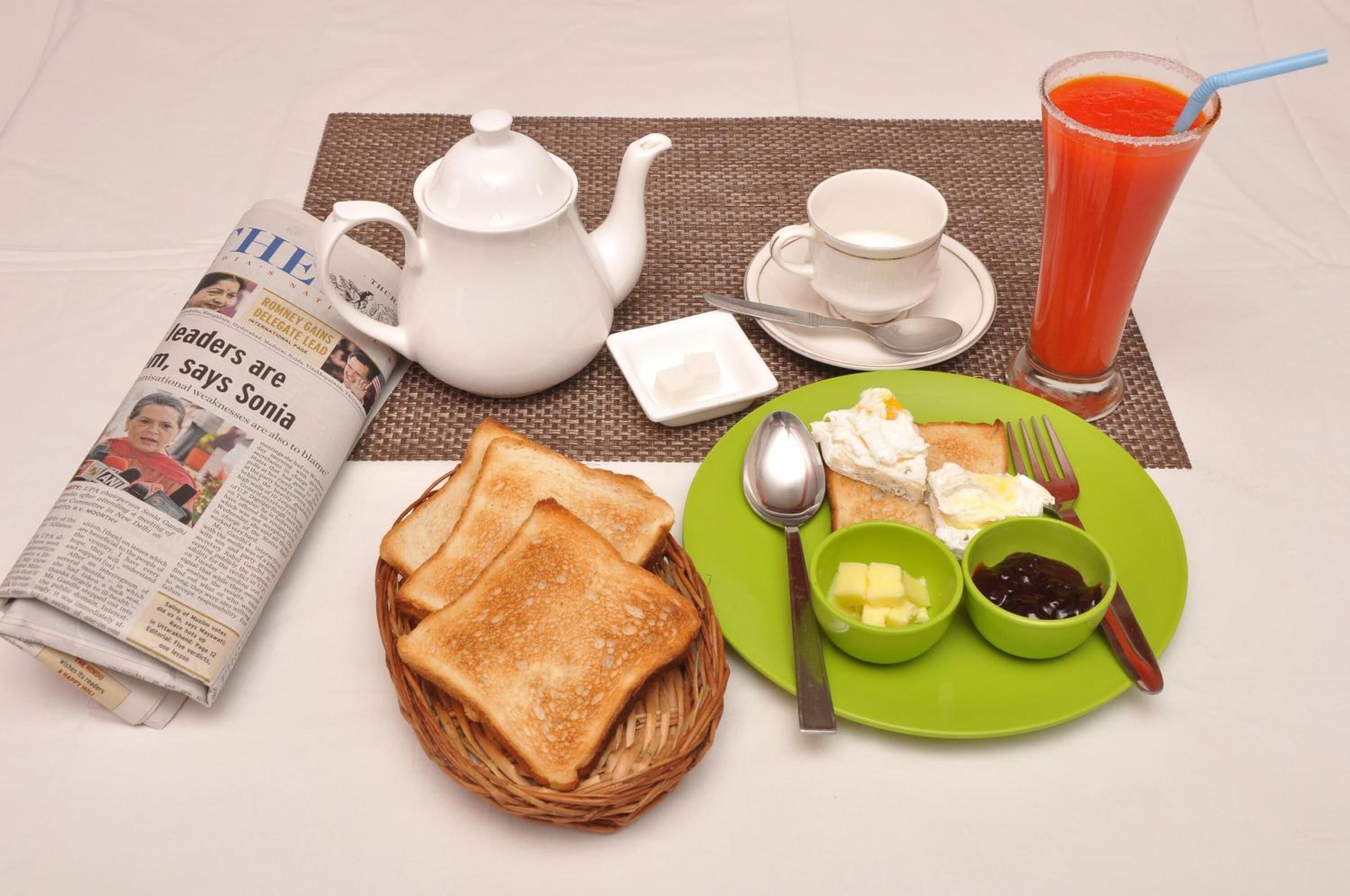 A breakfast setting with toast, butter, jam, a teapot, a cup, a saucer, a spoon, a fork, a newspaper, and a glass of juice.