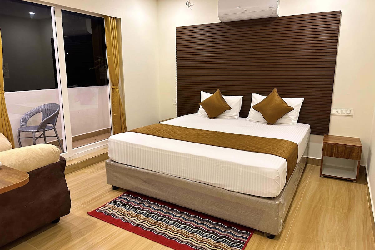 A hotel room with a large bed, a bedside table, a chair, a window with curtains, and an air conditioning unit on the wall.