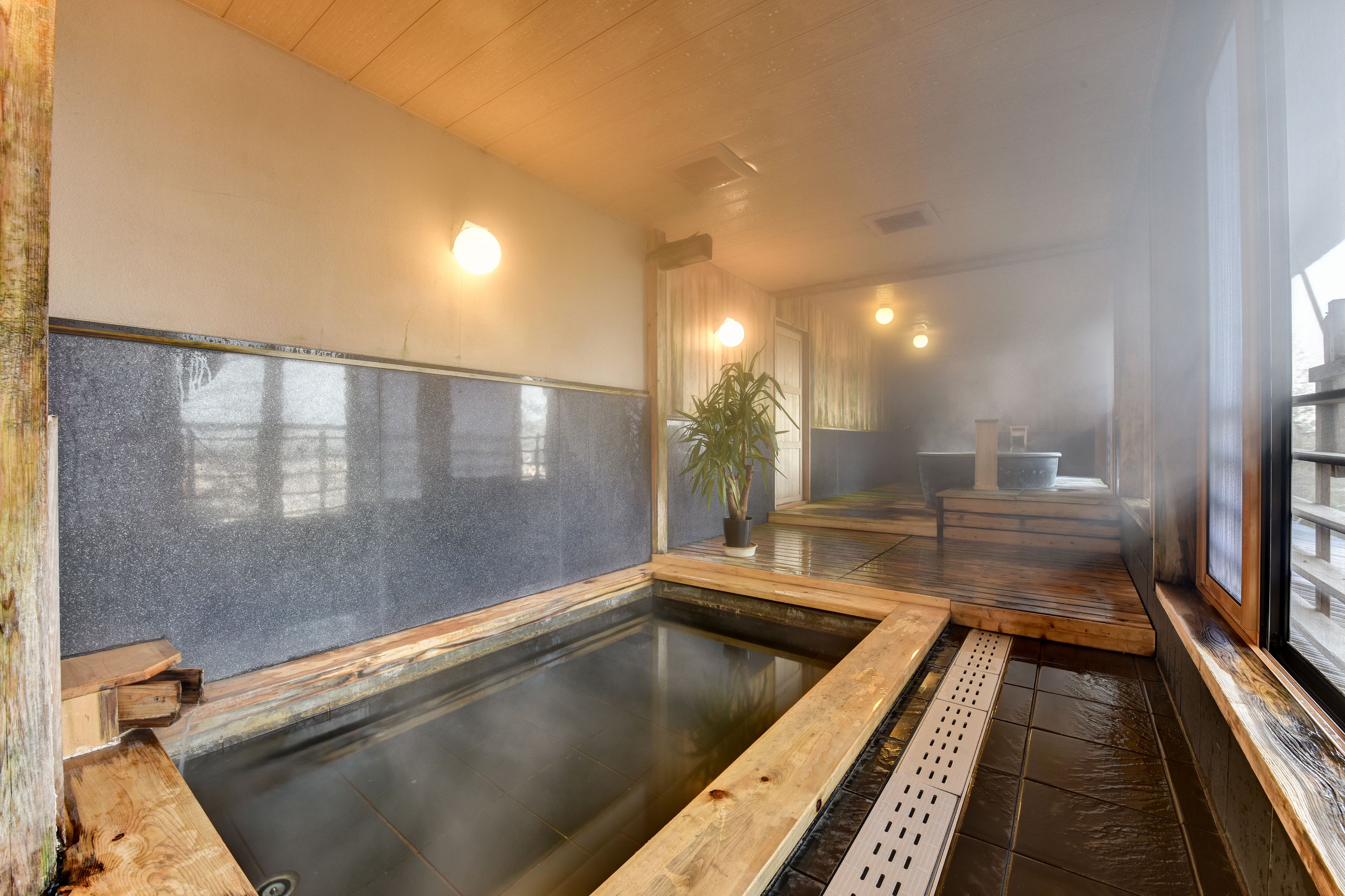 A traditional Japanese onsen with a wooden deck, a stone bath, and a view of the outside through a large window.