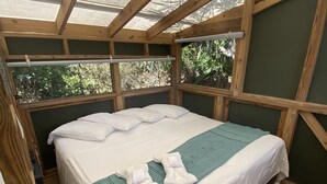 Un lit simple avec du linge de lit blanc et une couverture turquoise, des poutres en bois et une vue sur la verdure à l’extérieur.